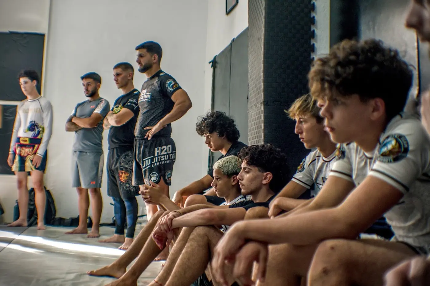 Entrenadores de pie con los brazos cruzados y alumnos jóvenes sentados junto a la pared, atentos a una explicación.