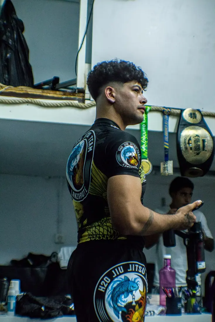 Amado de Jesús Aguilar Wade, entrenador principal, con la equipación del H2O Jiu Jitsu Team frente a medallas y cinturones de campeonato.