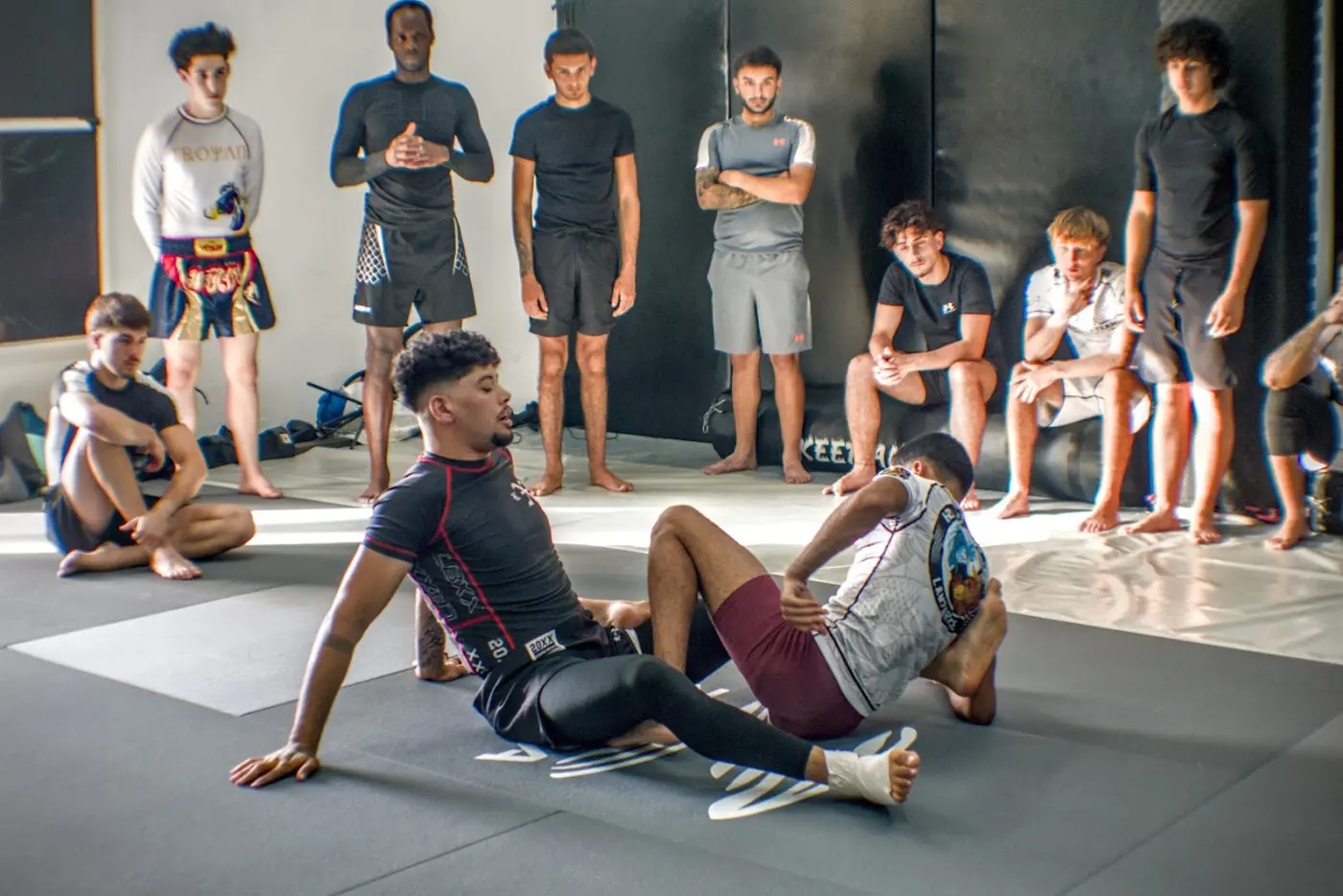 Dos deportistas demuestran una posición de grappling en el suelo mientras el grupo observa de pie.